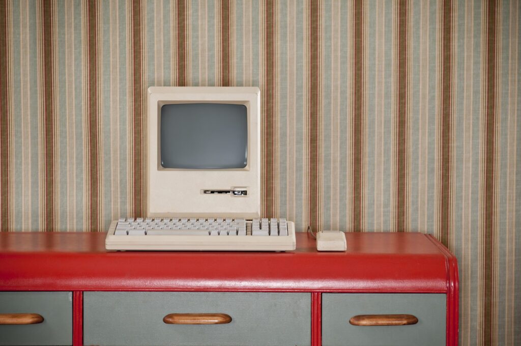 Old classic computer sitting on an art deco retro desk. The wall is covered in a wallpaper with a striped wallpaper.